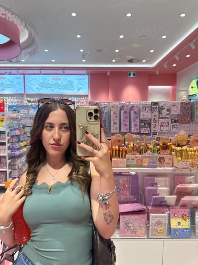 A woman takes a selfie in a store with pink and white decor. She's wearing a green shirt, rings, and bracelets. Sunglasses rest on her head. Her phone is visible, adorned with a sparkly case and decorative bow. The backdrop features shelves stocked with stationery items. The store appears to sell colorful, cutesy merchandise. The woman's shoulder tattoo is visible.