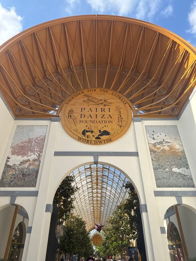 The image shows the entrance to Pairi Daiza Foundation in Cambron-Casteau, Belgium. A large, circular wooden sign with "TOGETHER FOR THE ANIMALS PAIRI DAIZA FOUNDATION WORLDWIDE" inscribed on it is prominently displayed. The sign features illustrations of a panda, a giraffe, an airplane, and a pagoda. Flanking the sign are two mosaic panels. The left panel depicts a tree with pink blossoms and traditional Asian architecture. The right panel shows a landscape with birds in flight above a tree and hills.

Above the entrance is a curved, wooden structure with a lattice-like design, supporting a portion of the building. The entrance itself is an arched walkway, covered by a glass and metal roof, extending into the distance and lined with tall trees. The sky above is blue with scattered white clouds. Below the arched entrance, people are visible walking through the pathway, suggesting a public space or attraction. The overall scene conveys an architectural entrance with artistic elements and a sense of greenery within a covered walkway.
