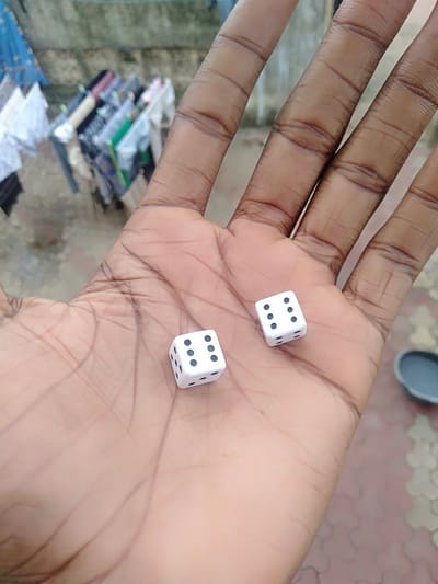 A human hand holds two white dice with black spots, showing the numbers "6" and "5". The background is blurry but appears to be an outdoor setting, with a tiled or patterned surface in the lower right and some undefined objects, possibly laundry, in the upper left. The image was taken in the city of Epe, Nigeria.