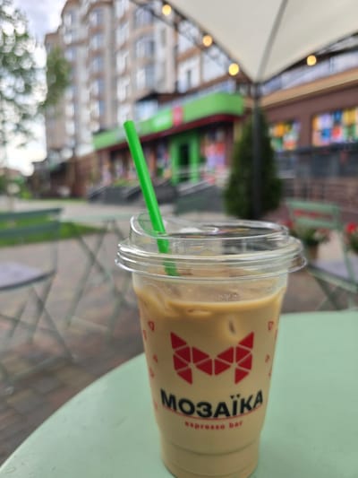 A close-up, side-view shot shows a to-go cup of iced coffee on a pale green surface. The cup is clear plastic with a fluted rim and a green plastic straw. The coffee is a light beige color and has ice cubes visible. The cup has a red geometric logo with triangles and the text "МОЗАЇКА espresso bar" in white and red lettering. The background is blurred, but appears to be an outdoor setting with buildings and trees. The lighting suggests it is daytime. The scene appears to be a casual outdoor cafe or street scene, possibly in Petropavlivska Borshchahivka, Ukraine, given the Cyrillic text on the cup. The overall mood is relaxed and ordinary.