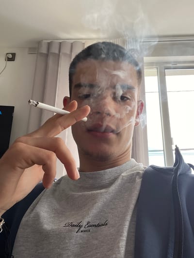 A man is depicted holding a lit cigarette in his right hand. Smoke is rising from the cigarette and partially obscures his face. He is wearing a grey t-shirt with the inscription "Daily Essentials MCMXCIX" on the right side of his chest and a navy jacket. The scene appears to be indoors, with a window and blinds visible in the background. An electrical outlet is visible on the wall behind the window. There is no evidence to suggest the location is Karavelovo, Bulgaria.
