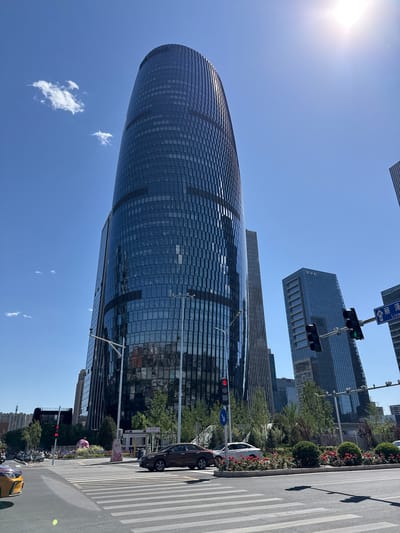 The image depicts a street scene in Fengtai District, China, featuring modern architecture and urban elements. A cylindrical skyscraper dominates the left side of the frame, its glass facade reflecting the clear blue sky with scattered clouds. To the right, a more conventional, rectangular skyscraper is visible. A street runs along the right edge of the image, complete with a pedestrian crosswalk, traffic lights (showing green and red), and a direction sign. Motor vehicles, including cars and motorbikes, are visible along the street, alongside a flowerbed or landscaping. A person holding a pink balloon is also seen on the sidewalk. The lighting suggests a sunny day, with the sun's glare visible in the upper-left corner.