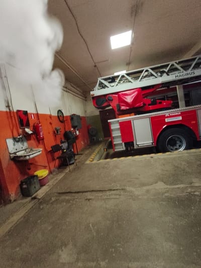 This image depicts the interior of a fire station in Tukums, Latvia. A red Magirus fire truck with a deployed ladder is prominently featured, partially obscuring the background. To the left of the truck, an orange wall is adorned with various items, including a sink, a fire extinguisher, and what appears to be a toolbox. Several buckets and other storage containers are visible on the floor. The ceiling of the station has fluorescent lighting. A significant amount of white smoke is emanating from the upper left portion of the image, obscuring details in that area. The overall environment suggests a functional workspace within the fire station.