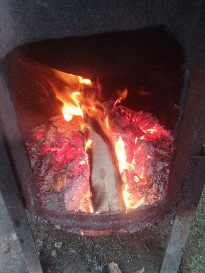 The image displays a contained outdoor fire in Nurli zamin, Uzbekistan. A dark, metallic, rectangular structure, possibly a brazier or fire pit, occupies the majority of the frame, viewed from above and slightly angled. Within this structure, active combustion is evident, featuring bright orange and yellow flames rising from several pieces of burning wood. A prominent, elongated log is centrally positioned, enveloped by flames and resting on a bed of glowing red embers and grey ash. The interior walls of the metal structure are dark and show signs of heat exposure. Directly below the base of the metal structure, a small portion of the ground is visible, characterized by sparse green grass and dark soil. No individuals are present in the scene.