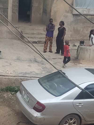 The image depicts a street scene in Alalubosa, Nigeria. The primary focus is a silver sedan parked on the right side of the frame. A group of four individuals are visible on the left side of the image, walking along a concrete walkway. The environment includes concrete building exteriors with weathered surfaces and a window. Power lines stretch across the scene, intersecting with the car.