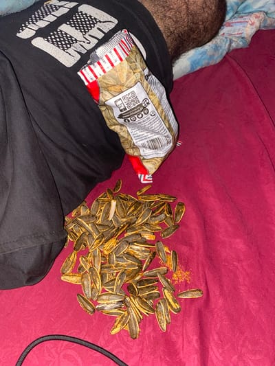 The image depicts a close-up, overhead view of a pile of roasted sunflower seeds spilled on a red fabric surface. A partially opened bag of sunflower seeds, with red and white stripes and visible QR codes and text in Arabic and French, is positioned at the top left of the frame. The bag appears to be a product of Algeria. A black t-shirt with a white graphic print is draped over the left side of the frame, partially obscuring the bag. A portion of a person's hairy leg is visible in the upper right background, suggesting the presence of a person. A black cable is visible in the lower left corner of the image. The overall scene appears to be indoors, possibly on a bed or couch, given the fabric texture and soft lighting. The specific location in Baraki, Algeria, is inferred from the product labeling on the bag.
