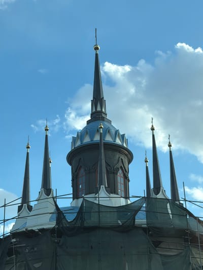 The image shows a low-angle view of the Church of the Vladimir Icon of the Mother of God in Bykovo, Russia. The camera points upwards towards the church's ornate spires and central dome, set against a background of a blue sky with scattered white clouds. Scaffolding and dark netting partially obscure sections of the church's exterior, indicating ongoing restoration or construction work. The church’s spires are dark and pointed, topped with gilded orbs. The architecture features intricate details and contrasting colors of blue, white, and dark grey.