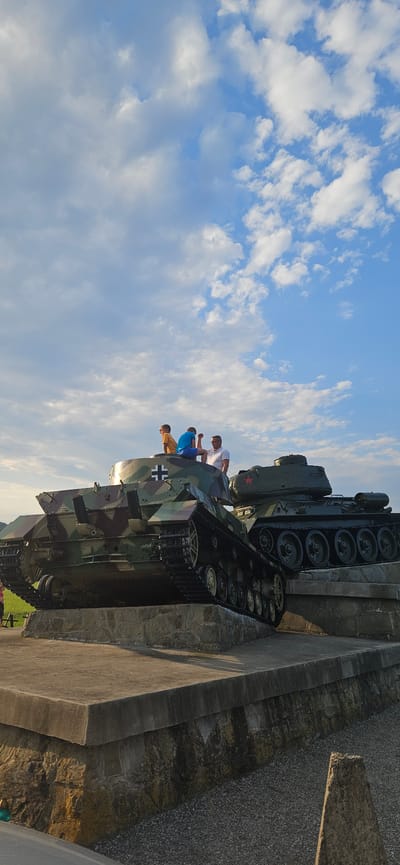 In Svidník, Slovakia, two tanks are displayed on a concrete and stone monument outdoors. The tank in the foreground, with camouflage paint and a white cross marking, is positioned at an angle. On top of this tank, three people are visible: two boys and a man. The boy on the left wears a yellow t-shirt and sits facing left. The boy in the middle wears a blue t-shirt and sits facing forward. The man on the right wears a white polo shirt and glasses and is seated. Behind the foreground tank, a second tank, painted green with a red star on its turret, is also on display. The background shows a blue sky with scattered white clouds and hints of a grassy field and trees to the far left.