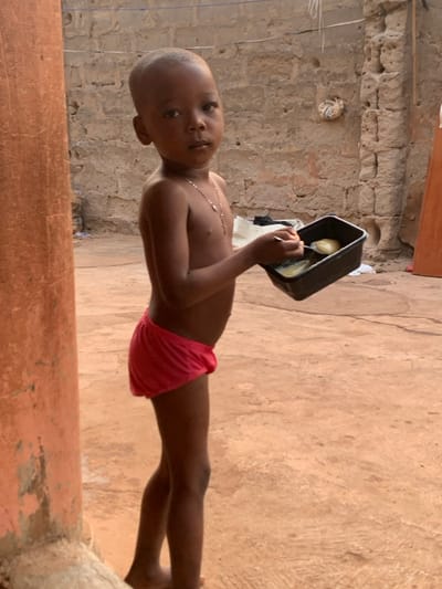 A young, shirtless child with close-cropped hair wears red shorts and a silver necklace. He stands on a brown, textured surface, holding a black container with food and a spoon. The child looks towards the camera. Behind him, a wall constructed of rough, tan-colored blocks is visible. In the background, a light-colored cloth item hangs. A weathered red-brown structure is in the foreground and partially obscures the lower portion of the image.