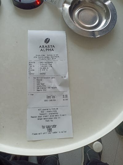 The image shows a receipt for a purchase at Arasta Alpha, a coffee shop located at Jalan Overste Isdiman, Ruko Widoro, Purwokerto Timur, 62114. The receipt details a purchase made on 13-04-2023 at 12:44, with Sales No: SMA01174452500428, Info: DAM.MKRA.45, and Purpose: 01 DIVE JN. The customer, D41FAN, purchased one Sea Salt Butterscotch Latte for 30,000 Indonesian Rupiah. The items included a regular shot of espresso, less sugar, Arasta Milk, 50% Arabica and 50% Robusta. The total cost was 30,000 Indonesian Rupiah, paid for by debit card. The receipt also includes information about the establishment's Wi-Fi and a queue number for the customer's order, 166. The image captures the receipt lying on a white circular table, with a pair of shoes visible at the bottom of the frame. The scene is a cafe in Purwokerto, Indonesia, where a customer has made a purchase and received their receipt.