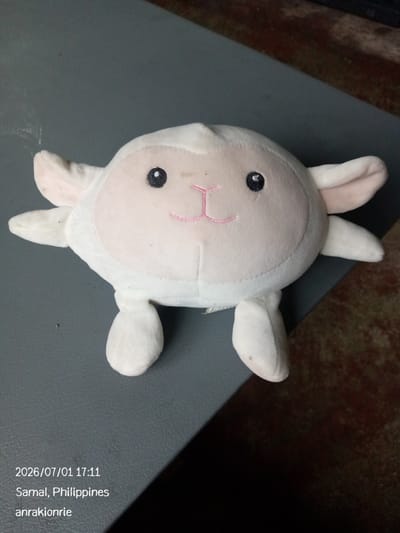 A white plush sheep toy with pink stitching for a mouth and black button eyes is lying on a grey surface. The toy is facing the viewer and appears to be in a seated position with its arms and legs spread out. The background is out of focus and shows a brown, textured surface and a dark area. The timestamp and location "2026/07/01 17:11 Samal, Philippines anrakionrie" are superimposed on the lower left corner of the image.