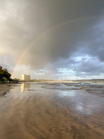 A wide-angle, low-angle shot captures a beach scene with a prominent rainbow arching across a cloudy sky. The foreground is dominated by wet sand, reflecting the sky and creating a mirror-like effect. The ocean is to the right, with gentle waves lapping at the shore. In the distance, a line of palm trees separates the beach from what appears to be a small town or resort. A tall, light-colored building stands out amidst the trees, its reflection visible on the wet sand. The sky is overcast with dramatic clouds, but patches of brighter light suggest either a break in the clouds or the sun is low on the horizon. A faint rainbow is visible on the left side of the frame, a subtle arc of color against the muted sky. The overall mood is serene and slightly dramatic due to the weather conditions. The time of day is likely late afternoon or early morning, given the soft light and the presence of a rainbow after or during rain. The location appears to be a tropical beach, with the building and vegetation suggesting a coastal resort area. There is no visible text in the image.