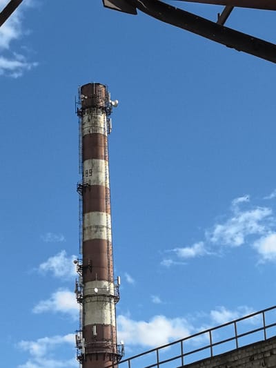The image depicts a tall, industrial smokestack against a clear blue sky with scattered white clouds. The smokestack is striped with alternating bands of reddish-brown and off-white, showing signs of weathering and age. Several metallic platforms and antennas are attached to the exterior of the smokestack at various levels, suggesting it may be used for telecommunications or other purposes in addition to its original industrial function. A metal ladder is also visible running along the side of the smokestack.
To the left of the smokestack, a number "89" is painted in black on one of the white bands. In the foreground and extending across the bottom of the image, there is a brick wall with a metal railing on top, indicating a boundary or walkway. Dark, angled metal structures are visible at the very top of the frame, possibly part of other industrial installations. A small, dark object, possibly a bird or an aircraft, is visible in the upper right quadrant of the sky.
The overall scene suggests an industrial area under a bright, possibly sunny day, given the clear blue sky and white clouds. The presence of the smokestack and the brick structures points to an urban or industrial setting. No people are visible in the frame. The mood is neutral, focusing on the architectural and environmental elements. The location context is Ivanovo, Russia.