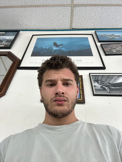 A man with short, curly brown hair and a beard poses for a photo. He wears a white t-shirt and is positioned against a wall adorned with various framed pictures. The pictures include images of airplanes and a nature poster. The wall is white, and a textured ceiling is visible in the upper left corner. The man's gaze is directed towards the camera.