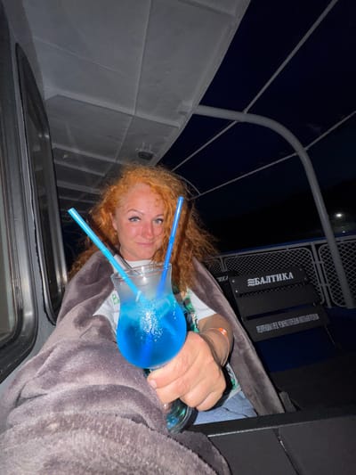 A medium shot captured at night shows a Caucasian woman with long, wavy red hair, wearing a light-colored shirt and blue jeans. She is wrapped in a dark grey blanket and holds a clear glass filled with a bright blue cocktail, complete with ice and two blue straws. She is looking directly at the camera with a slight smile. The background features seating on what appears to be a boat or ferry, with the name "БАЛТИКА" visible on one of the black seats. The environment suggests an outdoor setting at night, with dark skies and subtle lighting from the boat's interior. The woman is positioned on the left side of the frame, with the cocktail held prominently in her right hand. The scene is focused on the woman and her drink, with the background providing context of her location. The overall impression is one of relaxation and enjoyment.