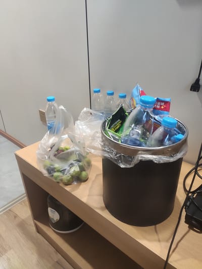 The image shows a black trash can filled with plastic bottles, snack bags, and some fresh produce. The trash can is located on a light-colored wooden shelf. Several plastic water bottles with blue caps are visible, some inside the trash can and others outside, partially covered in plastic bags. There are also a few snack bags with colorful packaging. A plastic bag filled with green fruit, possibly grapes, is also present. In the background, a white wall and a black power cord are visible. The overall scene suggests a casual setting, possibly a desk or a workspace, where items are being discarded or stored. The lighting is moderate, and the image appears to be taken indoors. No people are visible in the image. There is some visible text on the snack bags, but it is not legible. The image is oriented vertically.