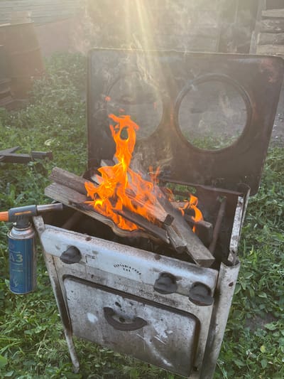 An outdoor scene in Syzran, Russia, features a rustic, old stove being used to burn wood. The stove, made of weathered metal, has two circular openings on its top surface and a door at the bottom. Several pieces of wood are stacked inside the stove and are engulfed in bright orange flames. A propane torch with a blue canister is positioned to the left of the stove, suggesting it was used to ignite the fire. The surrounding environment is grassy with some foliage and appears to be in a yard or rural area. In the background, there are large, dark cylindrical objects and a wooden structure. Sunlight streams down from the upper right, creating a bright lens flare and casting a warm glow over the scene. There are no people visible in the image.