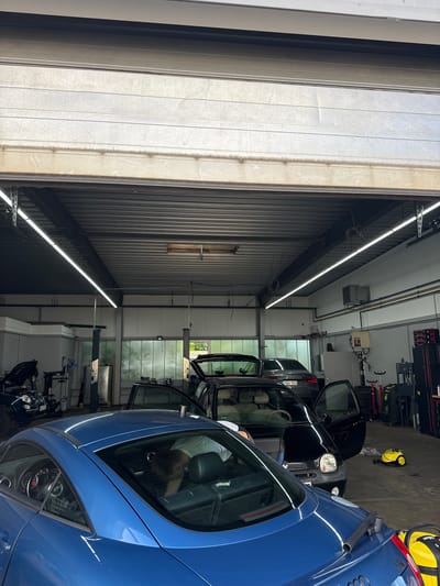 The image shows the interior of a car repair shop in Oberschleißheim, Germany. A large overhead door is partially open, revealing the workshop area. The shop is equipped with fluorescent lighting, car lifts, and various tools and equipment. Several cars are visible, including a blue sports car in the foreground, a black convertible, and a dark gray sedan. One person, partially obscured by the blue car, appears to be working on the interior of the vehicle. A yellow cleaning machine is also present on the floor. The environment appears to be functional and organized, consistent with a professional automotive service facility.