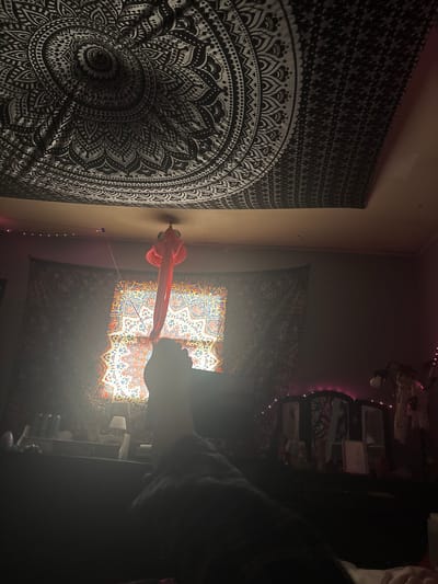 The image depicts an interior room in the City of Binghamton, United States of America, captured from a low, reclining perspective. A human left foot and lower leg, partially covered in dark fabric, occupy the lower-center foreground. The upper portion of the ceiling is draped with a black and white mandala-patterned tapestry. Directly below, a brightly illuminated window, covered by a colorful tapestry featuring red, blue, orange, and white mandala-like patterns, serves as the primary light source, creating significant lens flare. A bundle of reddish-orange fabric hangs from the ceiling in front of the window. The room's walls are adorned with dark, patterned tapestries. To the left of the window, a dark surface, possibly a dresser, holds various indistinct objects, including a small lamp. To the right, a dark dressing table with a three-panel mirror is visible, draped with pink string lights. Additional small string lights are present along the upper left wall. The overall lighting is dim, contrasting with the intense light emanating from the window. No other individuals are present.