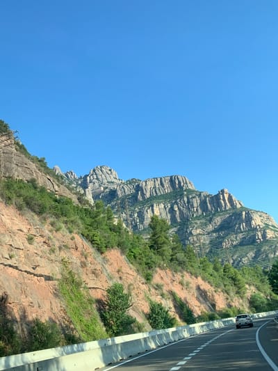 The image depicts a road curving through a mountainous landscape under a clear blue sky. The road, paved with asphalt and marked with white dashed lines, is bordered by a concrete barrier. A white SUV is visible driving away from the viewer on the right side of the road. The left side of the road features a steep, reddish-brown cliff face covered in scattered green vegetation, including trees and shrubs. Metal netting is visible on the cliff face, likely to prevent rockfalls. In the background, a range of jagged, layered mountains dominates the scene, with more trees and vegetation clinging to their slopes. Several tall, metal utility poles with wires are scattered throughout the landscape, suggesting the presence of power lines. The overall impression is of a scenic drive through a natural, rugged environment. The location is specified as Esparreguera, Spain.