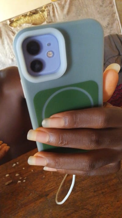A close-up, vertical shot shows a person's hand holding a smartphone with a light blue case and a circular green design on the back. The phone's camera module is visible, featuring two dark lenses and a smaller flash. The hand has long, light-colored fingernails and dark skin. The background is blurred, with what appears to be a wooden surface in the foreground and a textured fabric in the background. The image is taken in an indoor setting, likely a home. There is no specific indication of a location within the city of Donga, Nigeria.