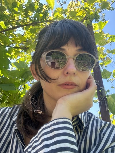 A woman with dark hair and bangs is looking at the camera, wearing sunglasses and a striped shirt. She is resting her chin on her hand. The background consists of green leaves and a blue sky, suggesting an outdoor setting. The image appears to be a selfie. The location is Zvonarevka, Russia.