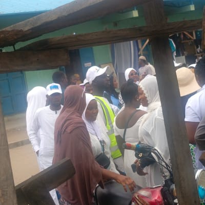 A gathering of people in Gudi, Nigeria is observed through the frame of a rough wooden structure. The group largely consists of individuals wearing white garments, many also wearing white head coverings. One person wears a brown hijab. A man in a white cap and a bright yellow reflective vest stands out. A red motorcycle is parked near the foreground. Blue painted doors are visible in the background.