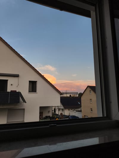 A twilight view through a window looks out onto a residential street in Pegnitz, Germany. The sky is a gradient of pale blue to soft pink and orange, with a large, illuminated cloud formation in the center. Several houses are visible, featuring white and yellow stucco facades and dark tiled roofs. One house on the left has solar panels on its roof and a rolling shutter over a window. A blue car is parked in front of a garage on the right. The foreground of the image is dark, showing the windowsill and window frame, suggesting the photo was taken from inside a building.