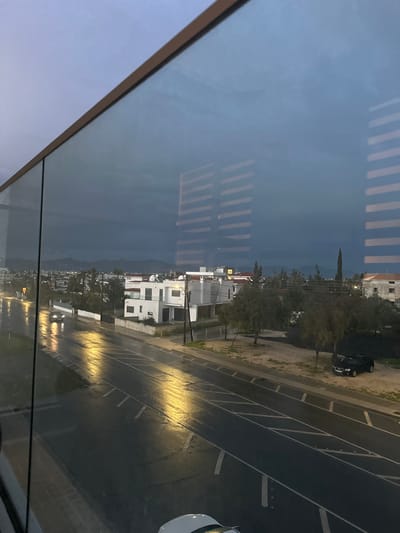 The image depicts an elevated view of a residential and commercial area in Latsia, Cyprus, under overcast and wet conditions, likely during dusk or dawn. The foreground is dominated by a clear glass barrier with a brown frame at the top, which reflects horizontal window blinds prominently across the upper two-thirds of the image.
Below the reflections, a wet asphalt road, indicating recent rain, stretches horizontally across the midground. The road surface is highly reflective, showing elongated yellowish streaks from artificial street lighting on the left side. Road markings, including broken white lines and diagonal hatching, are visible. One light-colored sedan with illuminated headlights is visible on the left side of the road, moving away from the viewer. A dark-colored SUV is parked on an unpaved shoulder on the right side of the road, adjacent to sparse trees and scrub vegetation.
Beyond the road, modern white multi-story buildings with flat roofs and visible solar water heaters are clustered on the left. Utility poles and overhead lines are present throughout the urban landscape. In the background, distant mountains are silhouetted against a heavily overcast, dark grey sky. The lower front portion of a white vehicle is partially visible at the bottom center, obscured by the glass barrier.