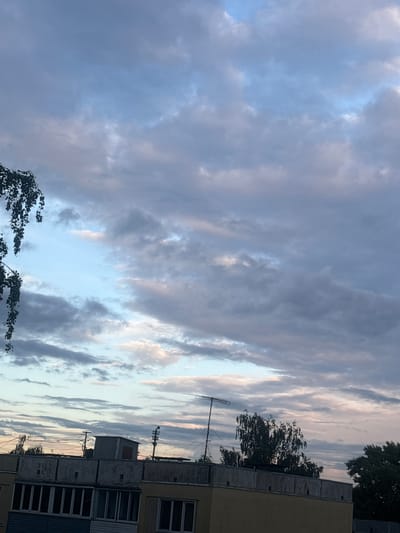 This is a vertical, eye-level shot of a sky filled with dramatic clouds. The clouds are a mix of dark grays and lighter whites and pinks, suggesting either an approaching storm or the aftermath of one, possibly at dawn or dusk. The lighting is soft and diffused.

In the foreground, the edge of a multi-story building is visible on the right side of the frame. It appears to be an older building with a textured, light yellow facade and multiple windows. Some of these windows have dark frames and appear to be covered with blinds or shutters. The building's concrete balconies are also visible, showing signs of wear and tear.

To the left of the building, there is a cluster of trees with dark green foliage. Their branches are partially obscuring the sky and the building. One tree in the lower left corner has branches that are bending and appear to be in motion, suggesting a gentle breeze.

A metallic antenna is mounted on a pole in front of the building, with several wires extending from it. In the middle ground, another smaller structure, possibly an air conditioning unit or a small shed, is visible on top of the building.

The overall impression is one of a calm, albeit slightly moody, evening or morning sky over a residential urban area. The lack of people or any specific activity suggests a moment captured in time, focusing on the natural beauty of the sky juxtaposed with the built environment. The time of day is likely either early morning or late evening due to the soft, diffused light and the colors in the clouds. The location context of Kirov, Russia, indicates an urban setting in that region. No visible text is present in the image.