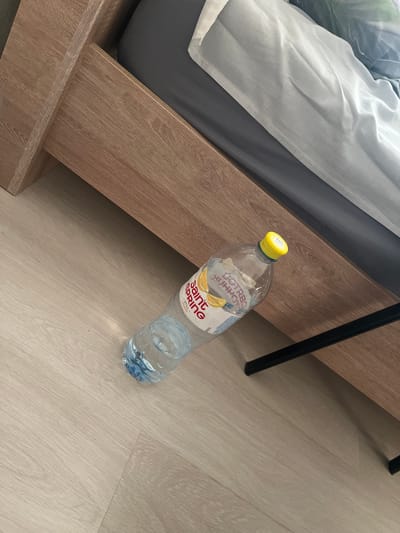 A partially filled plastic bottle of "Saint Spring" brand still lemon-flavored water lies on its side on a light-colored laminate floor. The bottle has a yellow cap and a white label featuring images of lemon slices. The bottle is positioned in front of a wooden bed frame, with the bed itself covered in gray and white bedding, partially visible. The scene appears to be indoors, likely in a bedroom, and does not provide any specific clues about its location within the city of Sorochinsk, Russia.