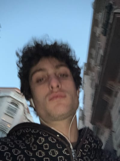 This image is a close-up, low-angle selfie of an adult male in an urban setting, consistent with the city of Vincennes, France. The male subject, approximately in his mid-20s, has dark, curly hair and light stubble on his jawline and chin. He is wearing a dark-colored, zippered jacket or hoodie with a repeating pattern of light-colored, stylized geometric shapes. White wired earbuds are inserted into both ears, with the wires visible extending downwards. His gaze is directed forward, directly at the camera, with a neutral facial expression. The sky above is a clear, light blue. In the blurred background, architectural elements are visible: a light-colored, multi-story building with a visible arched window is present on the upper left, while a darker, textured building facade is partially visible on the upper right. The background elements appear out of focus compared to the subject. The scene is illuminated by natural, even light.