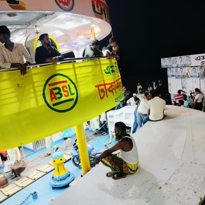 The image captures a vibrant night scene on a large vessel, likely a ferry or launch, in Bhola, Bangladesh. Numerous people are visible across multiple levels of the boat. On an upper deck, several men lean over a railing, observing the surroundings, illuminated by the vessel's artificial lighting. One man in a white striped shirt and another with a beard in a dark shirt are prominent figures on this level.
Below, on a curved white section of the deck, a man with a beard, wearing a white tank top and a yellow and brown plaid lungi, squats barefoot, looking forward. Further back on the main deck, groups of men are standing and sitting, some engaged in conversation or simply waiting.
A large, bright yellow banner hangs from the upper deck, featuring a green circular logo with the red and green letters "ABSL." Red Bengali script is also visible on the banner, partially showing "ঢাকা" (Dhaka), suggesting a destination or route. The deck features blue flooring, a yellow and blue mooring cleat, and wooden planks. A blue motorcycle is partly visible near the squatting man. In the dark background, on a structure to the right, numerous small political posters are affixed, each displaying multiple portraits of men in suits, likely election candidates. The dark sky confirms it is nighttime, with the vessel's lights providing illumination. The overall scene depicts a typical busy atmosphere on a public transport boat in the region.