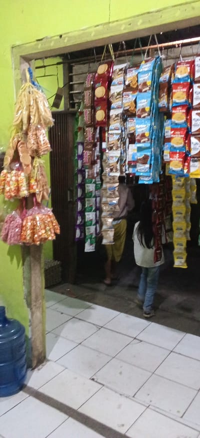 This image depicts the interior of a small convenience store, likely a warung, located in Banjaran, Indonesia. The walls are painted a bright lime green. A variety of packaged snacks, including cookies and crackers, are displayed hanging from hooks on a rack that spans across the doorway. Some of the snack packages are brightly colored, with prominent branding. To the left, a cluster of plastic-wrapped snacks, possibly fried snacks or crackers, hangs from a wooden post. In the background, a person wearing a light-colored shirt and blue jeans is seen walking away from the camera, while another person in a light brown shirt and yellow shorts is partially visible in the middle ground. A large blue water cooler jug is positioned on the floor to the left of the frame. The floor is tiled with white square tiles, some of which appear to be slightly uneven. The overall impression is that of a typical neighborhood shop in Indonesia.