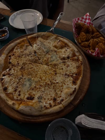 A pizza, sliced into eight pieces, sits on a wooden platter. The pizza is topped with cheese, exhibiting a golden-brown, speckled appearance. Next to the pizza, a bowl filled with fried food, possibly waffle fries or similar snack, is lined with a red-and-white checkered napkin. A drinking glass, silverware, and small sauce dish surround the pizza on the table. The scene is lit dimly, with a dark green tablecloth serving as the background.