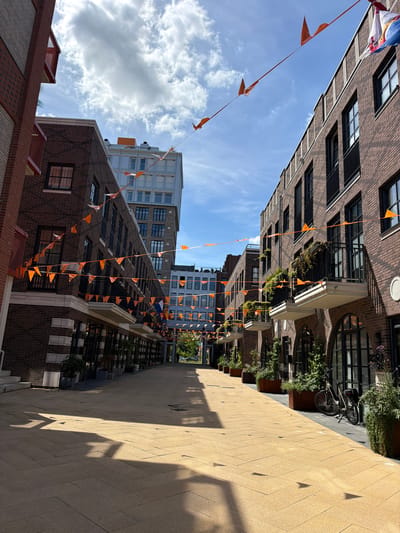 A street lined with modern brick buildings is decorated with orange triangular flags strung across the sky. The street is paved with yellow pavers laid in a herringbone pattern, and dappled sunlight casts shadows on the ground. Buildings on either side of the street have multiple stories, with balconies and large windows. Some windows have black frames. A bicycle is parked near a building on the right. The sky is blue with scattered white clouds. The scene is likely in Helmond, Netherlands, as indicated by the orange flags, which are a national color of the Netherlands and often used for national celebrations.