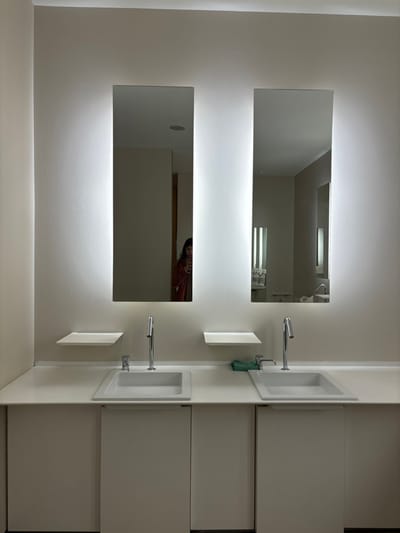 The image shows a bathroom with two sinks and two mirrors. The mirrors are rectangular and framed with bright, white lights. The sinks are also rectangular and white. The walls are white. Two silver faucets are visible, one for each sink. The image appears to be taken by a person standing in front of the sink. The reflection of the person's head and shoulders is visible in the lower mirror. The bathroom is located in Minato City, Japan. The coordinates (latitude: 35.662, longitude: 139.716) place the bathroom in a modern apartment building in the heart of Minato City. The bathroom is clean and well-lit. The environment is very quiet and still.