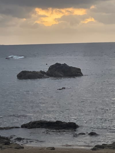 The image depicts a seascape with a cloudy sky. The water is dark and appears somewhat choppy, with several dark rocks jutting out from the surface. In the foreground, there is a sandy shore with more rocks. The sky is a mix of dark gray clouds and patches of warm, golden light, suggesting either sunrise or sunset. The scene appears to be a natural coastal environment. There are no identifiable landmarks or features that would specifically place this image within Las Palmas De Gran Canaria, Spain.