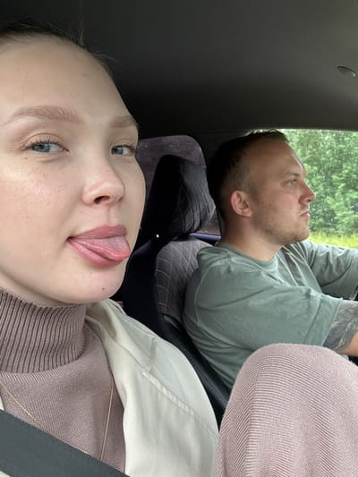 A woman with fair skin and blonde hair is in the foreground, sticking her tongue out playfully. She is wearing a light-colored jacket over a taupe turtleneck and a gold necklace. Beside her, a man with short brown hair and stubble is driving. He is wearing a green t-shirt and has a tattoo on his left arm. They are both inside a car, with the woman's face taking up most of the left side of the frame and the man's profile visible on the right. The background shows a blurred view of trees and possibly rain on the car window, suggesting they are on a road trip outside of a city. The interior of the car shows black leather seats and a dark headliner. The overall impression is a candid, lighthearted moment captured during a car journey. The specific location in the city of Novaya Kazmaska, Russia, cannot be determined from the visual information provided in the image.