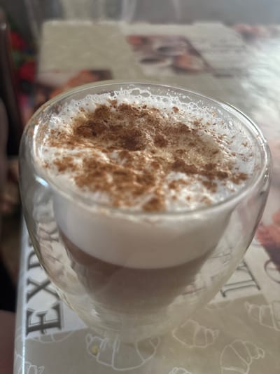A double-walled glass mug filled with a latte and topped with frothed milk and cinnamon powder sits on a table. The mug is clear glass with a visible layer of the latte beneath the foam. The table surface has a patterned tablecloth with images of croissants. The background is blurred, showing hints of a room and possibly another person. The scene is indoors.