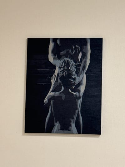 The image shows a painting of two figures in a dark, stylized depiction. The painting is square and has a dark background with lighter, greyish-blue figures rendered in a simplified, almost abstract style. The figures appear to be embracing or interacting closely, with one figure appearing to lift or support the other. The painting is hanging on a light-colored wall, possibly textured. The scene is indoors, and there is no indication of the city of Сосны, Russia.
