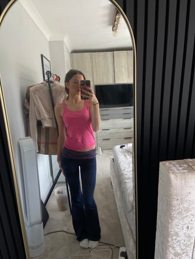 A young woman stands in a bedroom, taking a mirror selfie. She is wearing a pink tank top and dark blue flared pants. Her hair is pulled back, and she is wearing light makeup. The room is modern, with dark vertical paneling on one wall and a large mirror reflecting the space. A bed with a light-colored headboard is visible in the reflection, as is a dresser with drawers. Clothing is hanging on a rack to the left of the woman. The location is identified as Welwyn Hatfield, United Kingdom.