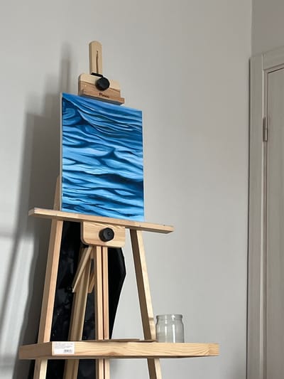 A painting of blue, wavy patterns is displayed on a wooden easel. The painting is rectangular and positioned horizontally. It appears to be an abstract piece with varying shades of blue. The easel is made of light-colored wood and is set up indoors, against a plain, light-colored wall.
The easel has several adjustable wooden pieces, and a black knob is visible on the left side, holding the painting in place. A glass jar is resting on the top right part of the easel. A black fabric or canvas is draped behind the painting on the easel. The overall lighting suggests an indoor setting, possibly a room or studio. The image is taken from a slightly low angle, looking up at the easel and painting.
There is some visible text on a white label attached to the right side of the easel. Additionally, the word "Pinax" is visible on the painting's support. The context provided indicates the location is Ильинское-Усово, Russia, though no specific visual cues within the image definitively confirm this location. The scene is static, with no action or event taking place beyond the display of the artwork. The mood is calm and focused on the art itself.