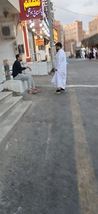 An outdoor urban scene in Makkah Al Mukarramah, Saudi Arabia, depicts two adult males interacting on a street at what appears to be late afternoon or early evening.
One man, seated on a low white concrete ledge adjacent to a building on the left, wears a dark long-sleeved shirt, light-colored trousers, and brown sandals. He has a beard and dark hair, and his right arm is raised in a gestural posture, indicating communication.
Facing him, the second man stands in the paved street, dressed in a white thobe and dark shoes. He also has a beard and dark hair, with his hands positioned low in front of his torso. Both individuals appear to be engaged in a conversation.
Behind the seated man, a light-colored multi-story building features a prominent red sign with illuminated Arabic text, reading "مطعم" (Restaurant) and "باكستاني هوٹل" (Pakistani Hotel/Restaurant). A smaller yellow sign with "RESTAURANT" is visible below. An air conditioning unit is mounted on the building's exterior.
The immediate foreground features two concrete steps leading from the street to the building. The street itself is paved asphalt with some faint markings. In the background, multiple multi-story buildings extend down the street, and blurred figures suggest additional pedestrian traffic. The sky above is a light grey-blue, indicative of dusk or an overcast day, complementing the active artificial lighting of the restaurant sign.