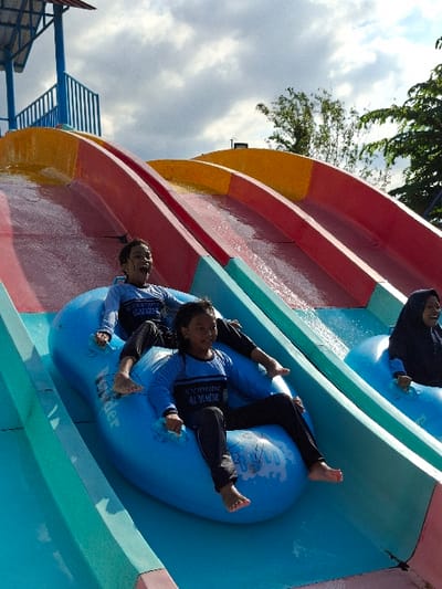 Two young girls are on a water slide. They are sitting in a blue inflatable ring, with the girl in the front wearing a black and blue long-sleeved shirt with white lettering on it. The girl in the back is wearing a blue long-sleeved shirt with white lettering. The girls are at a water park in Kecamatan Tulung, Indonesia. The park is located at approximately latitude: -7.592, longitude: 110.65. The slide is made of concrete and is painted in red, yellow, and blue. The slide has a slight curve to it. There is another girl sitting in a separate blue inflatable ring on another slide to the right of the image, but the girl is mostly cut off from the image. The water slide is surrounded by greenery, and there is a cloudy sky in the background.