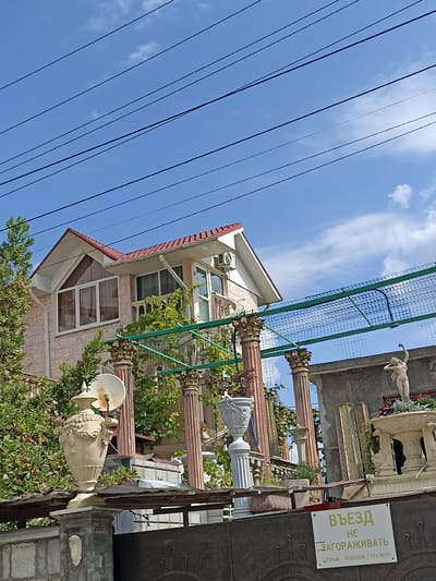 The image depicts a view of a building facade and its surroundings in Dagomys, Russia. A light pink, two-story house with a red tiled roof is visible, featuring white trim around the windows and a white gutter system. An air conditioning unit is mounted on the exterior wall. The foreground is occupied by a stone wall and a dark metal gate, with various decorative garden elements. These include large, ornate urns, classical-style pillars, and a statue of a female figure. A white sign with Russian text is attached to the wall near the gate. Green foliage from trees and vines is present throughout the scene, and multiple power lines are strung across the bright blue sky, which has a few scattered white clouds.