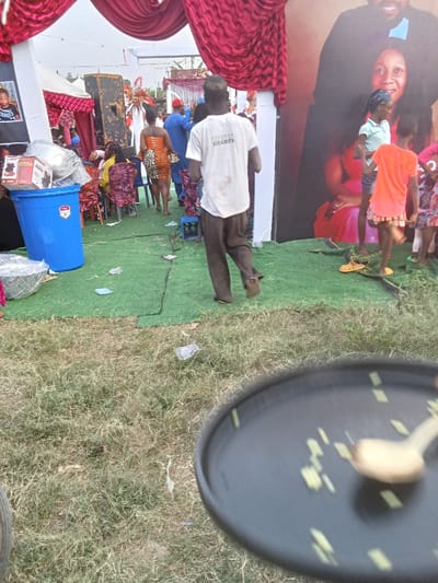 This image depicts an outdoor social gathering in Orie Uratta, Nigeria. The ground transitions from natural grass in the foreground to a spread of artificial green turf in the midground and background, both surfaces exhibiting scattered litter including paper and plastic.
In the blurred bottom right foreground, a dark, circular object resembling a tray or plate is partially visible, containing a light-colored wooden spoon and several small, pale food particles.
In the midground, a male individual is walking from left to right, seen from behind. He wears a soiled white t-shirt with the text "FOREVER HEARTS" printed on the back and dark trousers, with indistinct dark footwear. To his left, beneath a white tent structure draped with elaborate dark red fabric, are multiple attendees. A woman in an orange and black patterned dress is visible from behind, near a large dark speaker. A man in a blue shirt and red cap is positioned further back. A large blue plastic bin filled with packaged items, including boxes labeled "HOME PRO," is situated on the far left. To the right of the walking man, a large vertical banner displays a photographic portrait of an adult man and woman, with a child's image visible at the bottom. Several children are gathered near this banner, including a girl in a light blue top and another in an orange dress. A pair of yellow sandals rests on the artificial turf near the children.
The background shows additional white tent structures with matching red drapery and decorative elements, suggesting an organized event. The sky appears overcast.