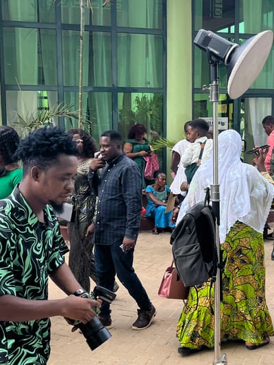 The image depicts an outdoor gathering in Madina, Ghana, featuring multiple individuals and professional photography equipment against a building with a light green facade and large, dark-framed windows.
In the left foreground, a male with dark, curly hair and a beard, wearing a black and light green patterned short-sleeved shirt, operates a camera with a telephoto lens, looking downwards. Behind him, a male in a dark plaid long-sleeved shirt and dark jeans walks towards the right, holding a white paper in his left hand and his right hand near his right ear, as if in conversation.
To the right of the central male, a silver light stand supports a black Godox AD600BM strobe light head fitted with a large white parabolic reflector. A black backpack hangs from the light stand. A female in a white hijab and a long, green, yellow, and brown patterned skirt stands to the right of the light stand, holding a mobile phone to her right ear.
A young child, dressed in a white shirt, green shorts, and suspenders, is held by an adult largely obscured by the light stand, positioned to the left of the hijab-wearing female. The child faces right.
In the mid-background, visible figures include: a female with large, curly dark hair and sunglasses, a female in a dark green dress inspecting a mobile phone, and an older female in a blue patterned dress seated on a low step. The ground is paved with reddish-brown bricks.