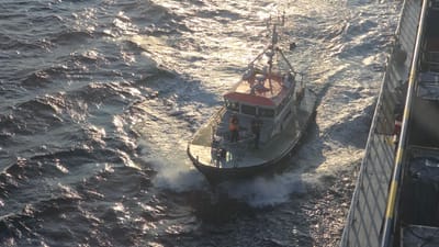 An aerial, long shot captures a pilot boat moving through choppy, sun-dappled water next to a larger vessel's railing. The pilot boat, a gray and red vessel with a cabin, has at least two individuals on its deck. One person, wearing an orange vest, stands near the front of the cabin, while another individual in dark clothing stands behind them. The water is dark blue and reflects the golden sunlight, creating a textured, dynamic surface. The railing of the larger vessel, partially visible on the right, is gray with yellow and dark blue markings and casts long shadows. The scene suggests movement and activity at sea, likely in a port or coastal area. The context of Aberdeen, United Kingdom, would place this scene in the North Sea, potentially near the city's active harbor.