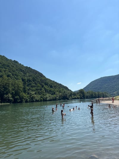 The image shows a sunny day at a body of water, likely a river or lake, with lush green hillsides rising from its banks. The water is clear and calm, with ripples on the surface. Several people are in the water, some swimming, some standing, and some appearing to be playing or relaxing. The far bank is lined with trees, and a rocky area is visible in the upper part of the image, where more people are gathered. The sky is a clear, bright blue, indicating good weather. The overall scene evokes a sense of leisure and enjoyment on a warm summer day.
